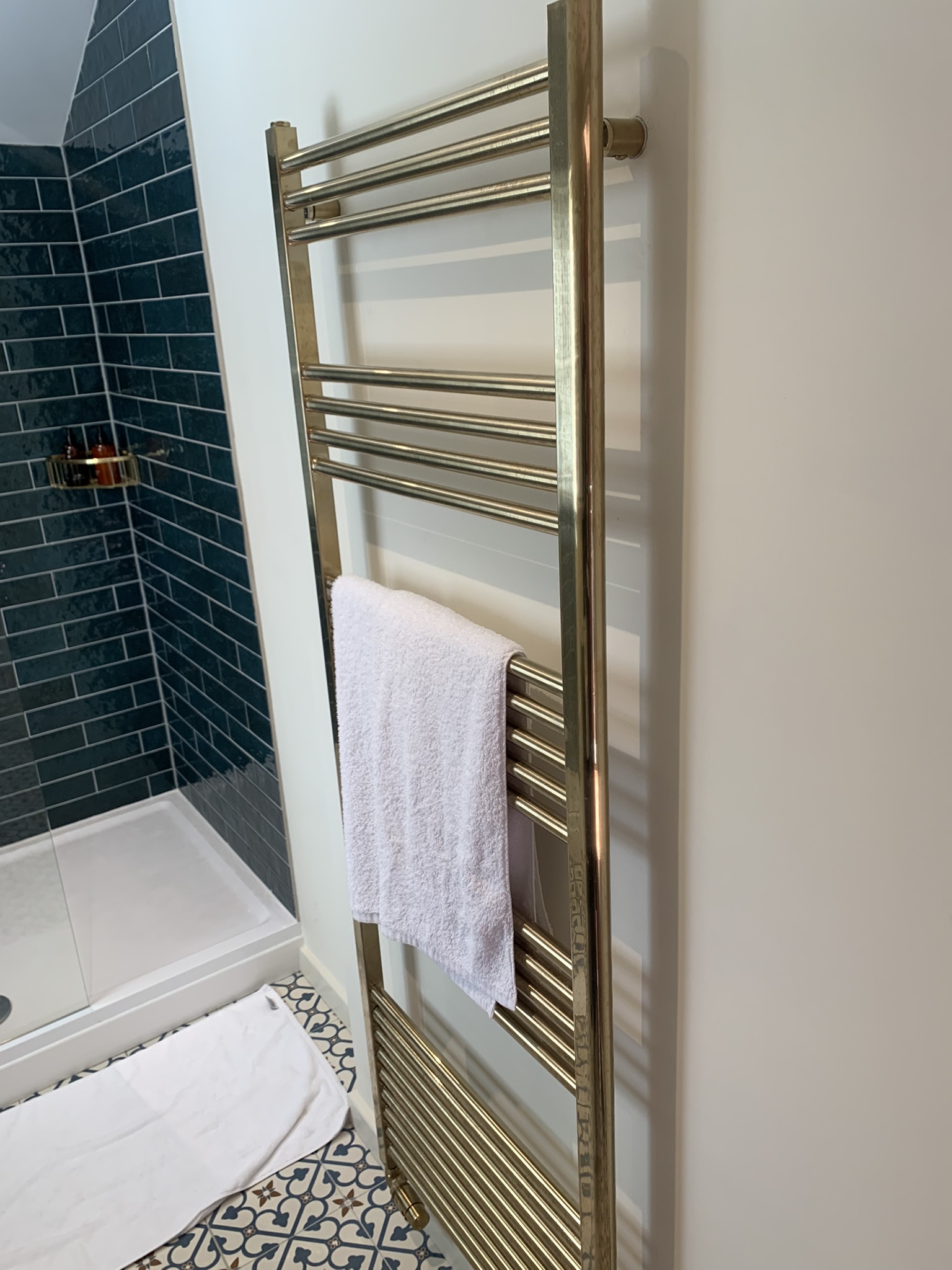 Brass heated towel rail beside a teal-tiled shower, with patterned floor tiles