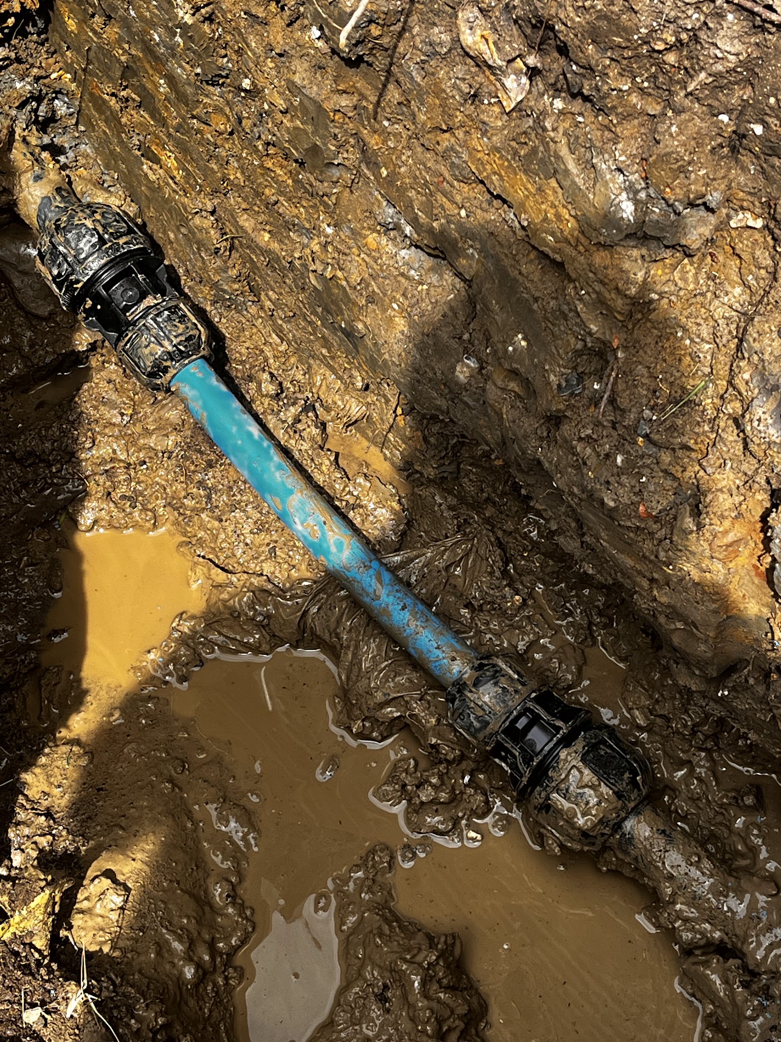 An excavated blue water pipe with new compression fittings in a muddy trench during a leak repair
