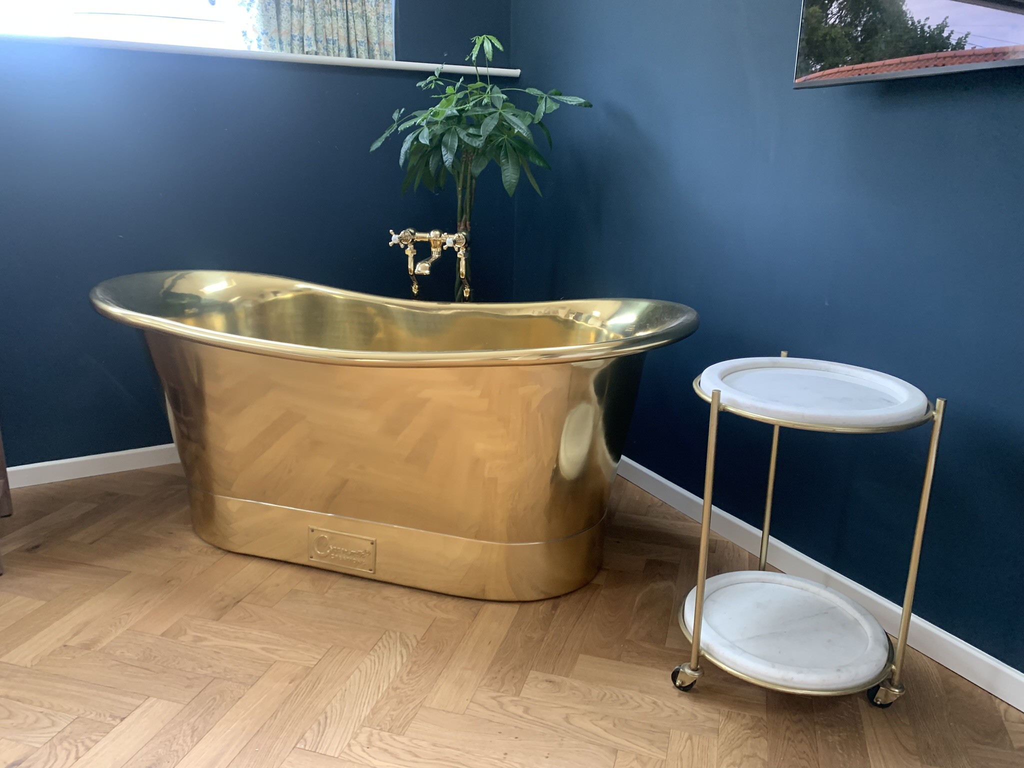 Polished brass slipper bath against a navy wall on a herringbone oak floor, fitted by Matt McBride